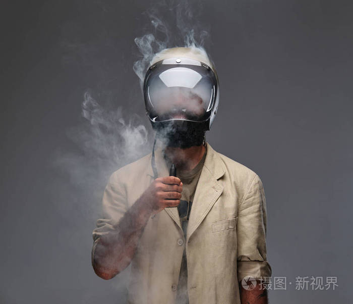 戴着摩托车头盔的长胡子的时髦人抽着电子香烟.在灰色背景下隔离