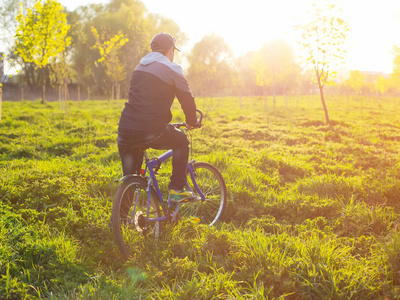 在日落期间在一条乡村公路穿过绿色的春天草地上骑自行车的年轻人