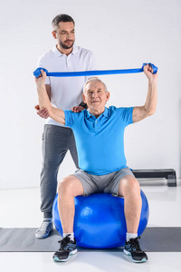 康复治疗师协助资深男士在健身球上用橡皮带运动