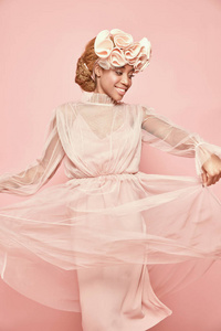 时尚肖像的迷人的微笑黑女人与轻头发和绿色的眼睛穿着粉粉红色薄纱礼服和大褶边箍和摆姿势。粉红色背景