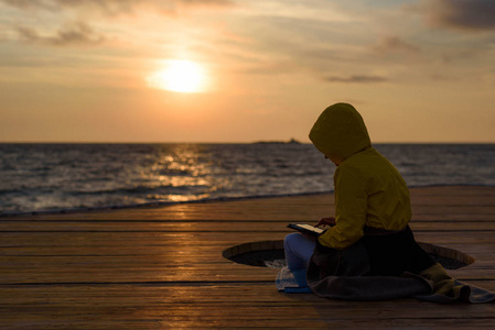 黄色外套的女孩剪影与毯子在木码头与平板电脑在日出的海洋背景