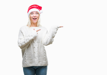 年轻美丽的金发女子戴着圣诞节帽子在孤立的背景惊讶和微笑的相机, 而用手和手指指着