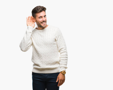 年轻英俊的男子穿着冬季毛衣在孤立的背景微笑与手超过耳朵听谣言或八卦的听证会。耳聋概念