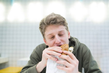一个年轻人在一家清淡的餐馆里咬一口美味的汉堡包。饥饿的学生吃汉堡包与乐趣