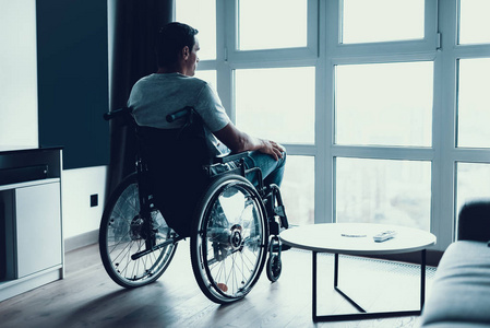 残疾人坐在轮椅上靠窗。严肃的悲伤白种人穿着休闲装, 在明亮的现代客厅或医院看大型全景