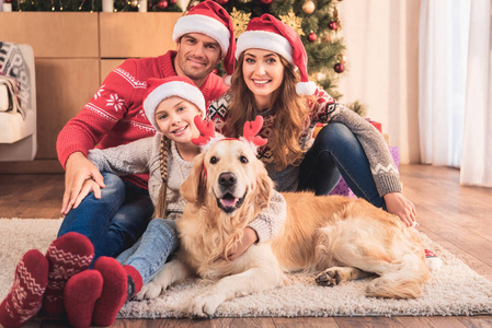 幸福的家庭在圣诞帽子与狗在鹿角坐在附近的圣诞树