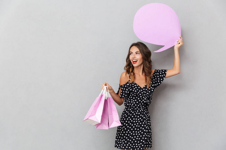 一个笑的年轻女孩的肖像在灰色背景下, 背着购物袋, 显示空的语音气泡