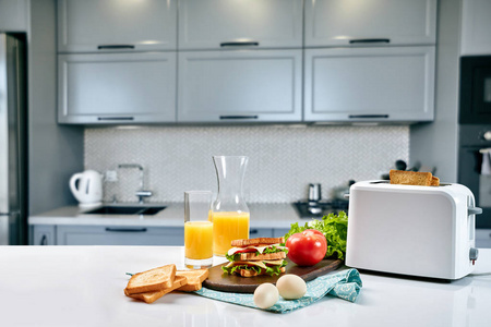 白色电动面包烤箱与橙汁与三明治在厨房桌上。在舒适的厨房里享用早餐