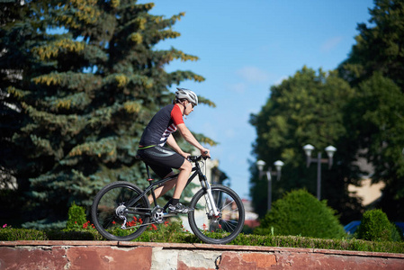 年轻人专业自行车骑自行车在街道边缘附近花坛和树在红色大厦前面