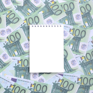 白色笔记本上的清洁页躺在一套绿色货币面额100欧元。很多钱形成无限堆