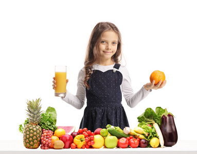 女孩拿着一杯果汁和一个橙色的桌子后面的水果和蔬菜孤立的白色背景