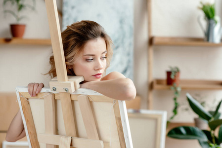 美丽的感性的年轻女子倚在画架和看着在艺术工作室