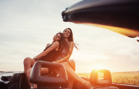 年轻的两个女人在拍照。女孩高兴地摆在黑色的汽车旁边的天空上的梦幻般的日落