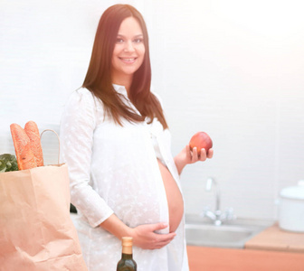 年轻的怀孕女子捧着一个苹果，站在厨房