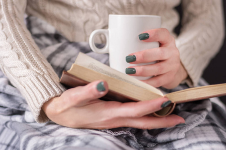 手里拿着书和一杯咖啡的女人穿着毛衣, 盖着一条复古的毯子。冬冷天气概念。特写, 选择性聚焦