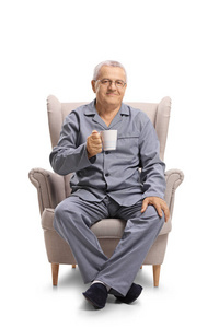 穿着睡衣的成熟男人坐在扶手椅上, 在白色背景下拿着一个杯子