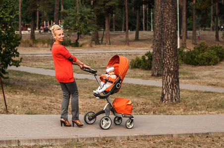 一个年轻的微笑的母亲带着一个橙色的婴儿推车与一个女婴在公园里