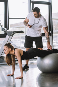 男性私人教练与定时器和年轻的运动妇女做俯卧撑健身球在健身房