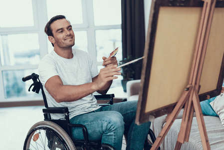 轮椅画中的残疾人。微笑英俊的快乐白种人坐在对面的黄鼠狼在客厅穿着休闲服, 手持画笔和调色板