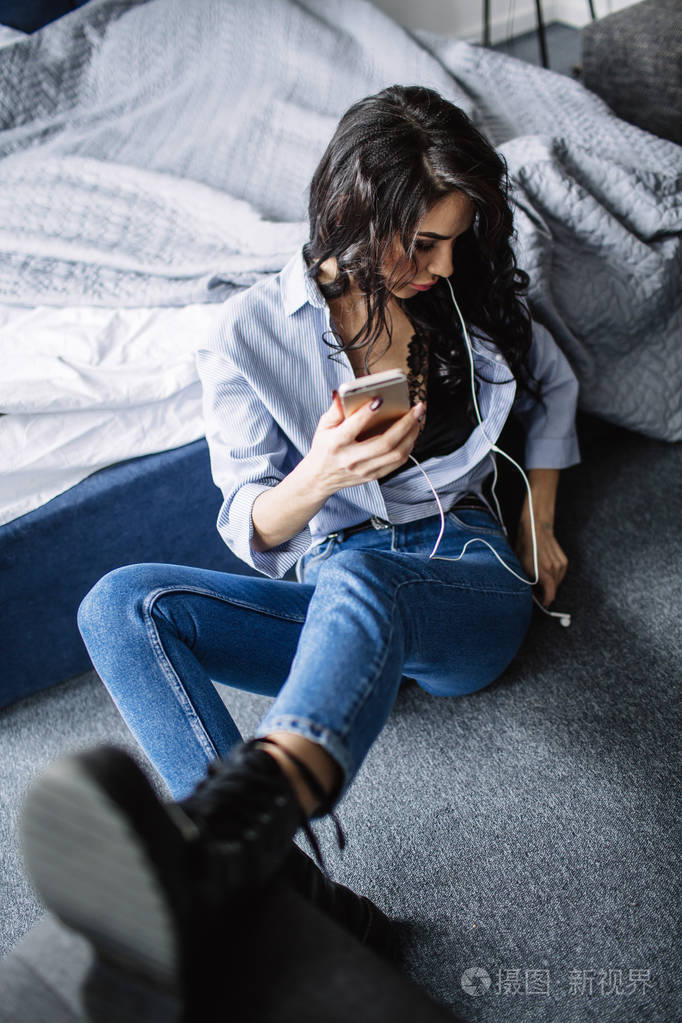一个漂亮的黑发女郎在沙发附近的房间里听音乐, 穿着衬衣和蓝色牛仔裤