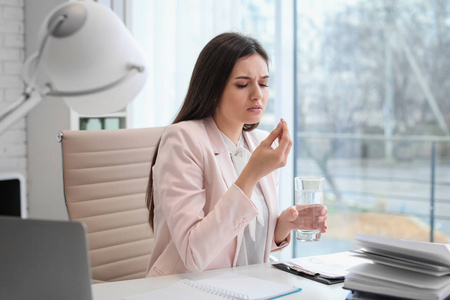 一位年轻女子坐在办公室的餐桌上吃药丸来对付头痛