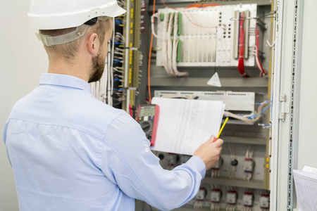 工程师在白色头盔阅读设计图纸反对电气工业面板。服务人员用控制器分析电气自动化控制箱中的电路