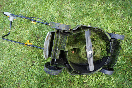 电动割草机躺在草地上, 轮子上下颠倒。在里面, 整个机制被湿切草堵塞