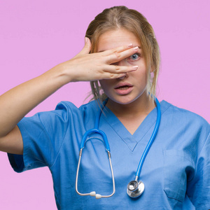 年轻的白人医生妇女穿外科医生制服在孤立的背景偷看在休克覆盖面和眼睛用手, 看通过手指尴尬的表情