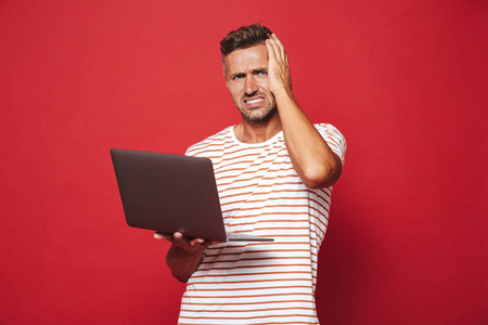 在条纹 t恤衫上的困惑年轻人的形象微笑和持有灰色笔记本电脑孤立的红色背景