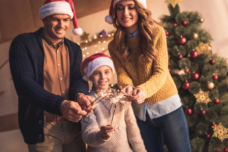 幸福的家庭与女儿在帽子举行烟花在家里与圣诞树