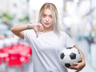 年轻的金发女子持有足球足球在孤立的背景与愤怒的脸, 否定的符号显示不喜欢拇指向下, 拒绝概念