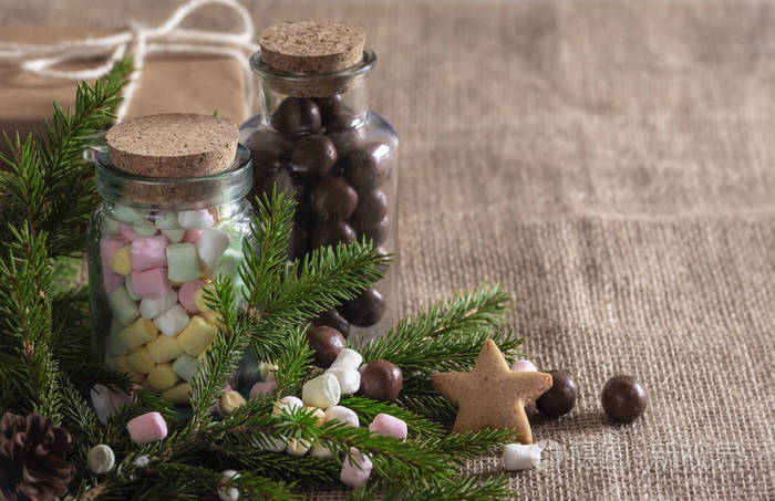 圣诞甜点背景五颜六色的迷你棉花糖和巧克力糖果在瓶子, 常青杉木树枝, 星形饼干, 黄麻织物上