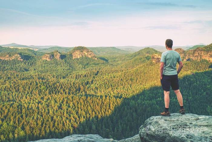 穿着户外衣服的高个男子独自站在岩石的顶峰在清晨的薄雾中观看丘陵