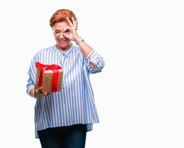 高级白种人妇女拿着礼物在孤立的背景与快乐的脸微笑做确定的标志与手在眼睛看着通过手指