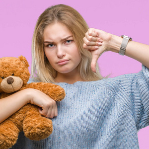 年轻的白种人妇女抱着可爱的泰迪熊在孤立的背景与愤怒的脸, 否定的符号显示不喜欢拇指向下, 拒绝概念