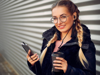一个时尚的年轻女孩的肖像与编织金发在小穗与圆玻璃与化妆听耳机在她的手机音乐和喝热饮料在条纹铁背景
