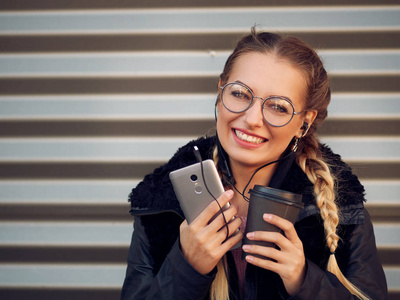 一个时尚的年轻女孩的肖像与编织金发在小穗与圆玻璃与化妆听耳机在她的手机音乐和喝热饮料在条纹铁背景