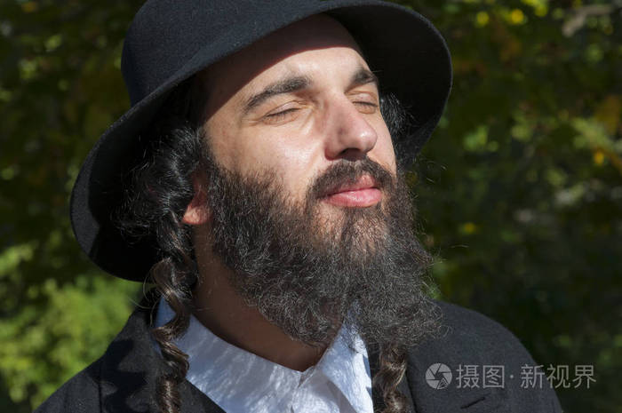 户外阳光灿烂的年轻传统正统犹太男子与眼睛关闭与黑色胡子, 帽子和