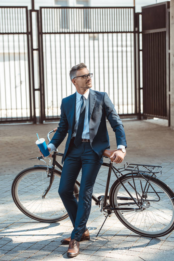 英俊的微笑的商人拿着纸杯, 坐在自行车上的街道上