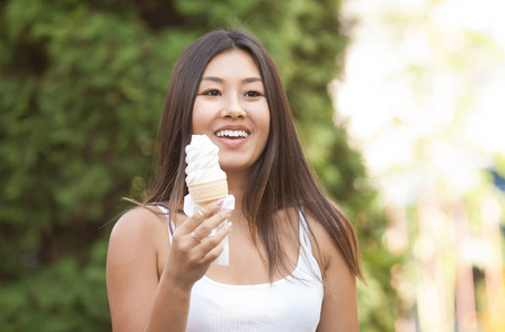 愉快的年轻的微笑亚洲褐发女郎妇女吃冰淇淋在绿色的树背景在公园