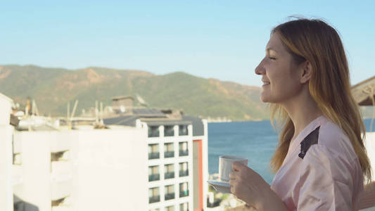 美丽的女孩浴袍喝香咖啡, 而站在她的房间阳台上的酒店