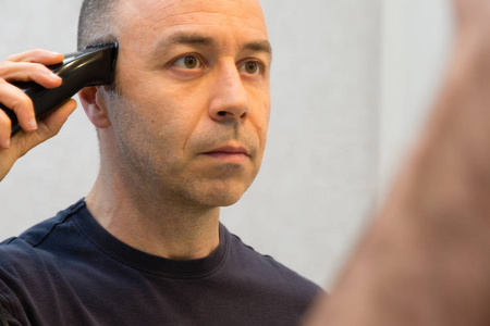 黑色衬衫光头男子剃光他的头发, 在镜子前的电动剃须刀照片