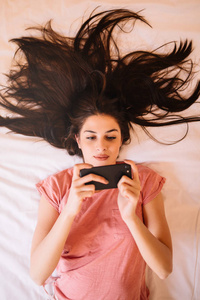 迷人的黑发人躺在床上, 使用手机, 上网冲浪, 发短信或玩游戏。上角视图