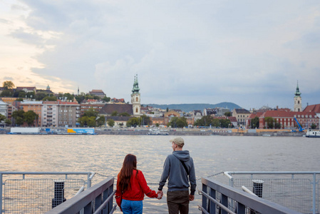 背后的迷人情侣在爱是牵手和欣赏多瑙河的景观在布达佩斯, 匈牙利在日落期间