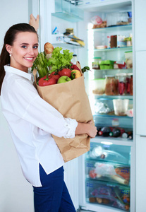 年轻女子捧着蔬菜杂货店购物袋。站在厨房里。在厨房里看着相机的女人