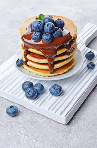 美味的早餐。在灰色的混凝土背景下, 自制的蓝莓和融化的巧克力在白色木制板材上的煎饼