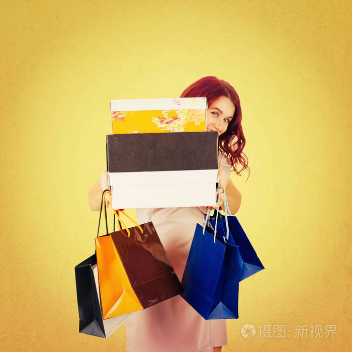 一个顽皮的年轻女子躲在购物箱后面, 手里拿着许多纸袋作为节日礼物, 在白色背景下被隔离。