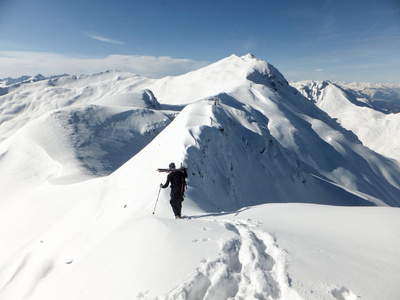 男子背部滑雪者背着滑雪板, 当他沿着一个狭窄的和裸露的积雪山脊在他的途中, 在山顶上导航