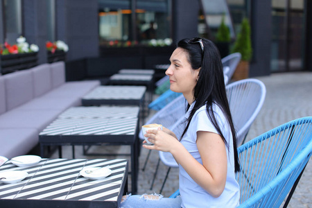 迷人的黑发妇女喝咖啡在咖啡馆和坐在蓝色椅子上