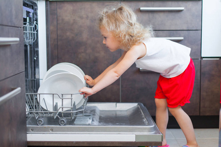 可爱的金发小孩姑娘的帮助在厨房里把印版划出洗碗碟机
